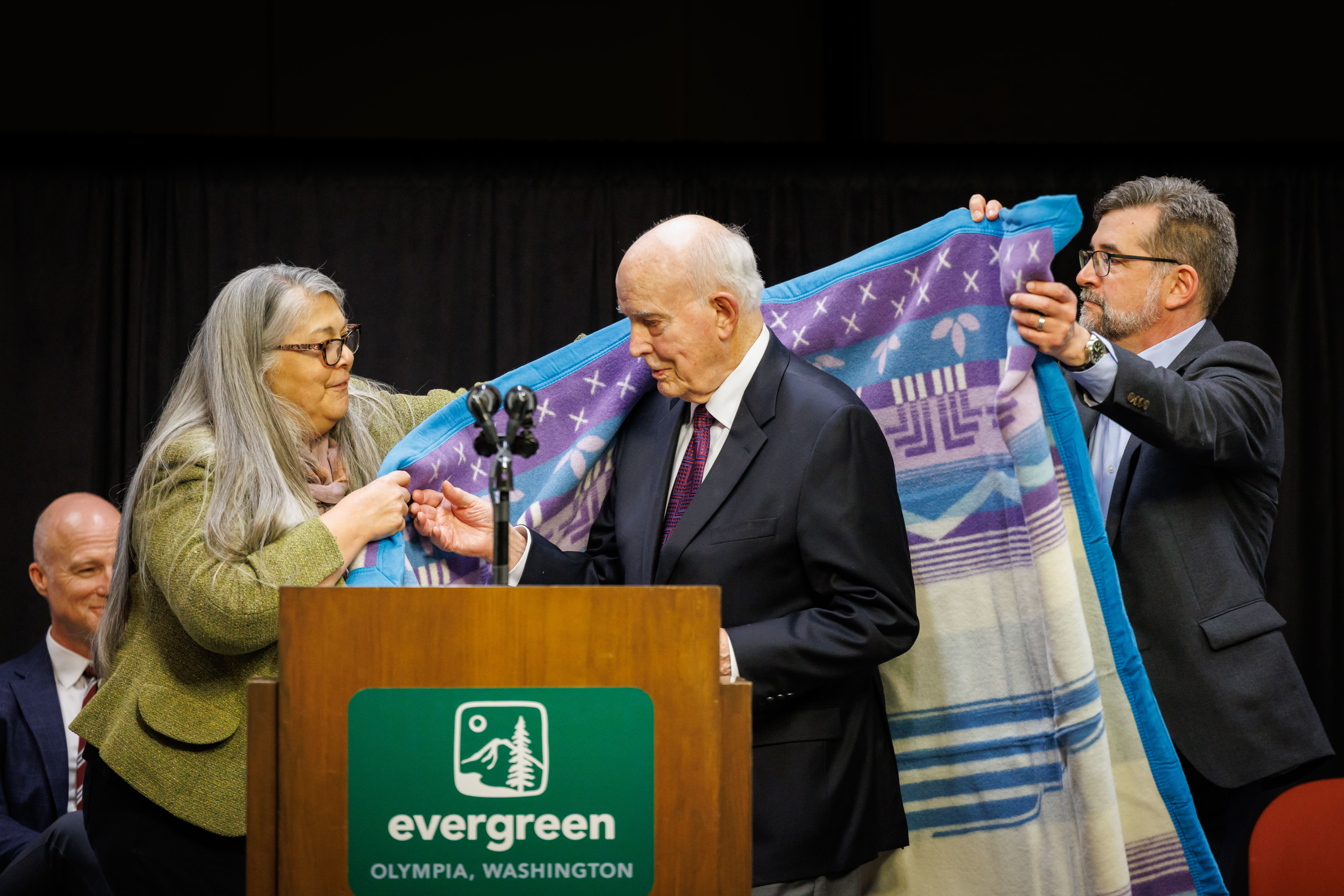 Daniel J. Evans being honored with a blanket by Evergreen President, John Carmichael and Director of the House of Welcome, Laura VerMeulen