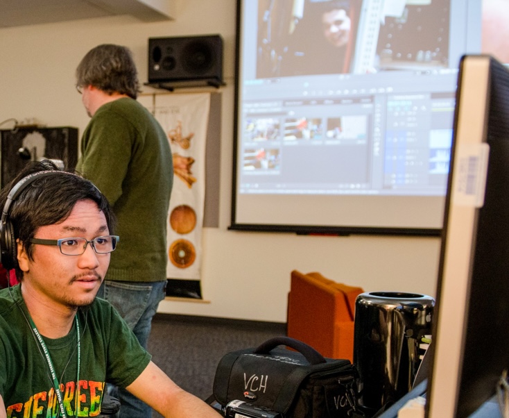 Student with glasses and mustache sits at a computer, while a video editing program is projected on the wall behind him