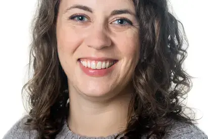 a photo of a white woman with curly brown hair and a grey sweater on, she is smiling at the camera