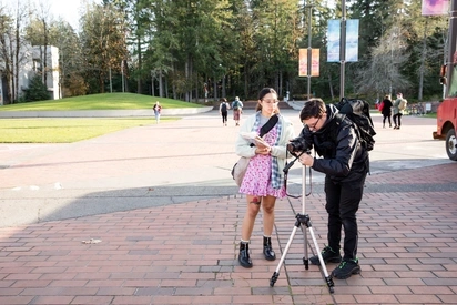 Two students work behind a camera on the Evergreen campus. One holds a clipboard and the other adjusts the camera.