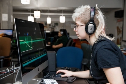 Blonde student wearing headphones, editing audio files on a Mac Computer
