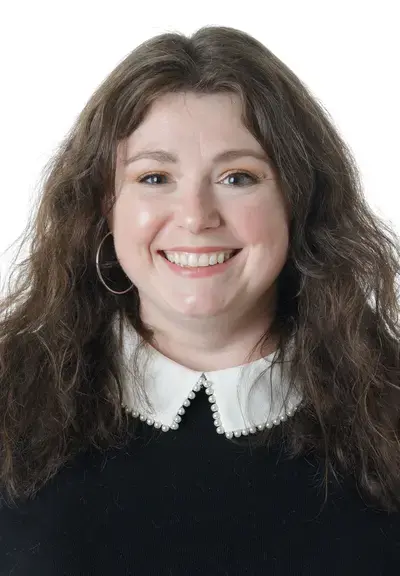 a white woman with long brown wavy hair smiling a the camera, she is wearing a dark sweater with a white collar coming out the top