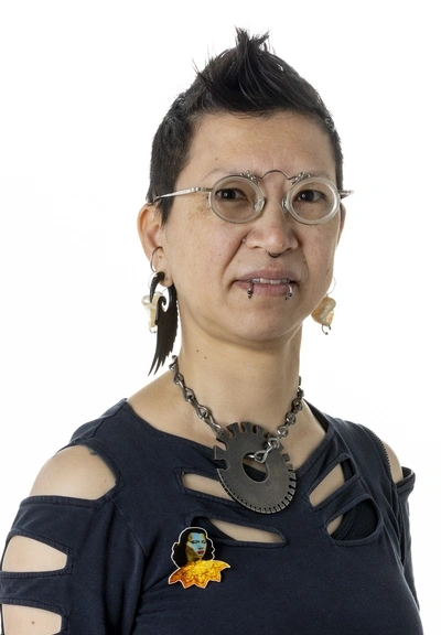 Headshot of person with dark, short spikey hair and snake bite lip piercings, wearing glasses, large earings, a large necklace. an a colorful pin.