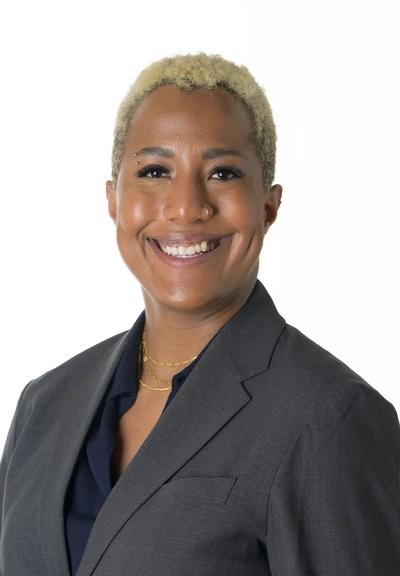 Headshot of a person with cropped blond hair, a big smile and wearing a grey blazer over a black button down shirt