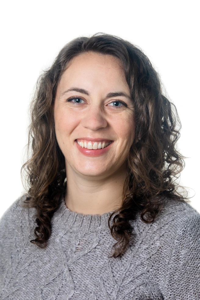 a photo of a white woman with curly brown hair and a grey sweater on, she is smiling at the camera