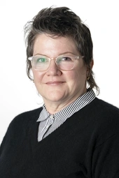 Person with short dark hair wearing glasses and a black sweater over a black and white striped buttun up shirt