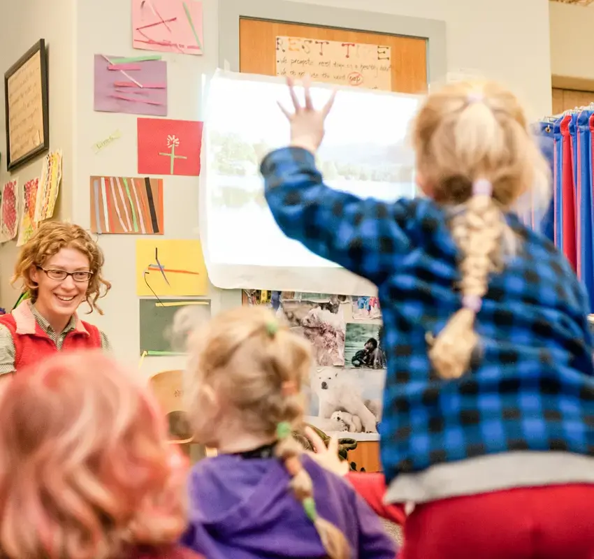 Children in a classroom. in the foreground a child with their back to the camera has their hand raised, the teacher in in the background