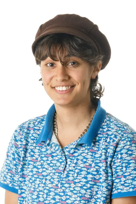 A person with medium legnth brown hair wearing a blue shirt with spots and a brown hat