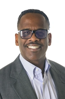 headshot of a person with short dark curly hair wearing dark rimmed glasses and a grey sports coat over a light grey button up shirt